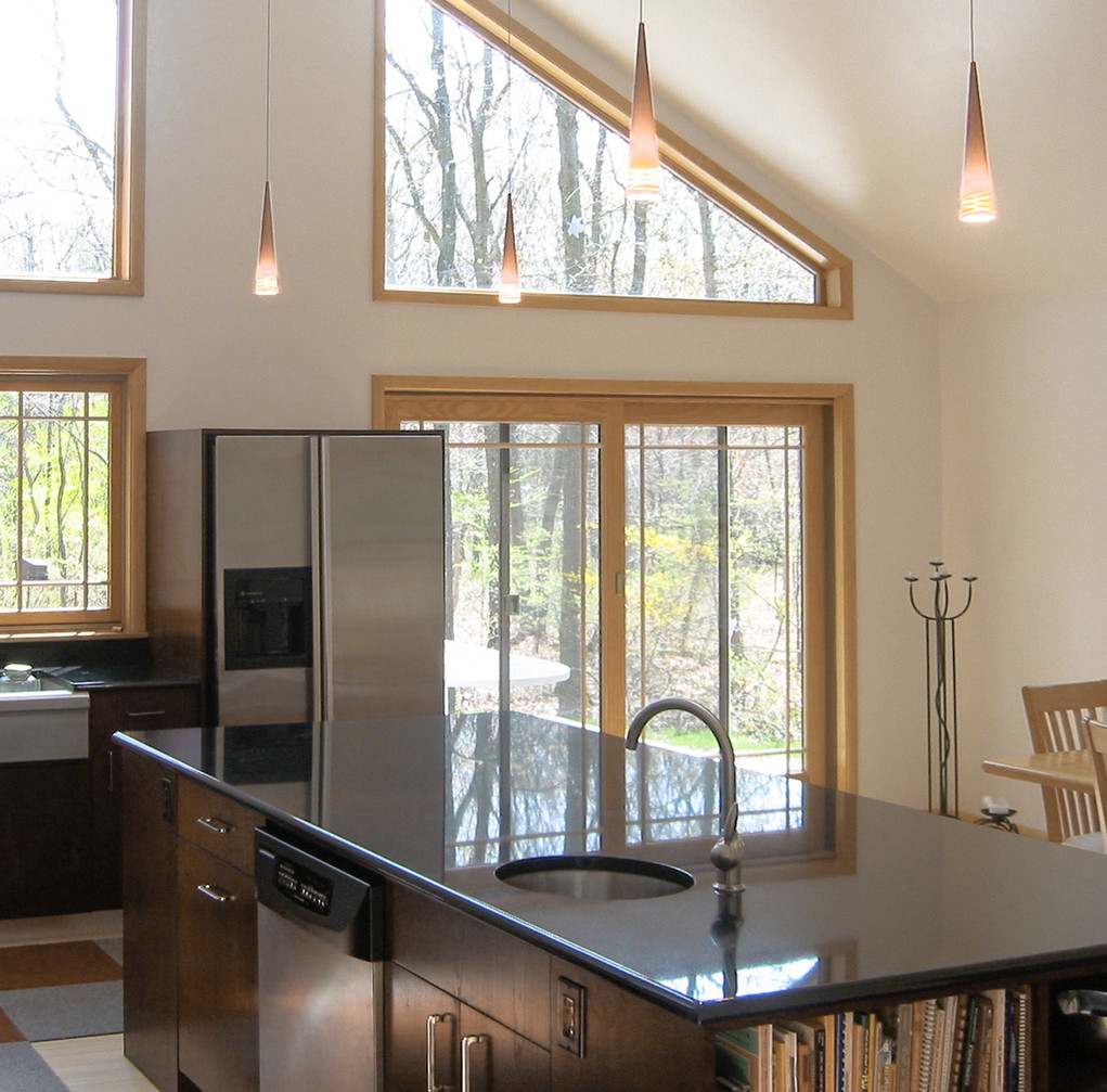 A modern redeveloped kitchen featuring a furnished kitchen island bar with a sleek black countertop. The space is well-lit with hanging cone-shaped pendant lights, and large windows offer a view of the natural surroundings. The kitchen includes stainless steel appliances, a sink integrated into the island, and wooden cabinetry with a touch of natural light streaming in from the windows.